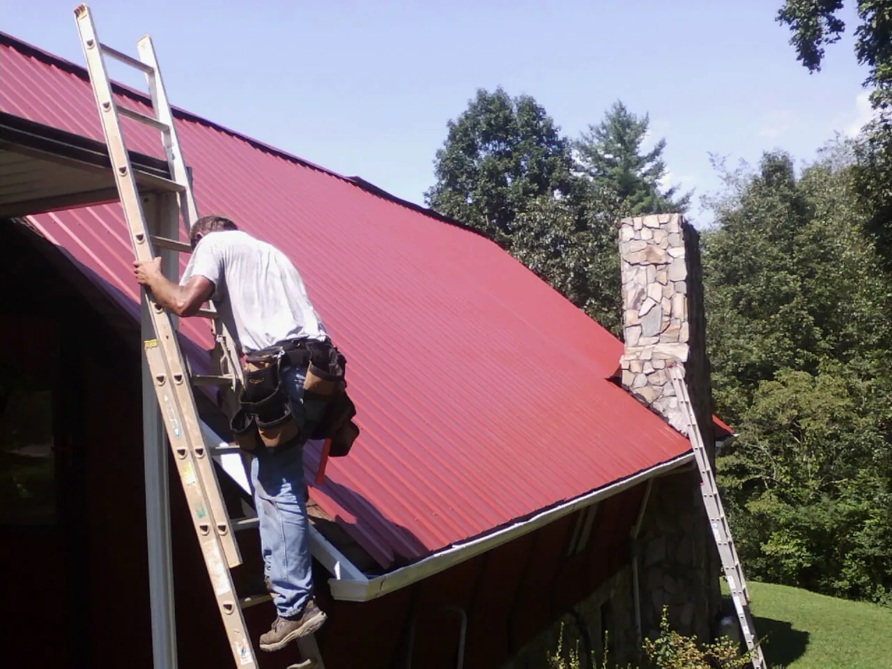Roofer on a ladder beside a red metal roof and stone chimney — Soffit & Fascia Repair by Roofing Brothers in Holladay